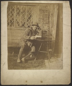 [Seated man with hat reading book at table outside house]