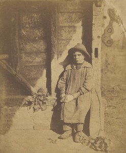 [Young boy in hat and smock seated outside peeling turnips]